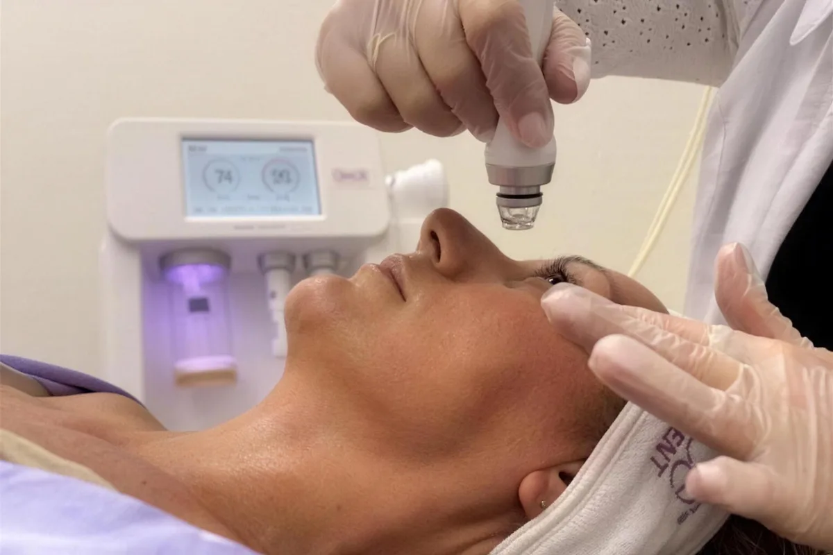 A practitioner guides a facial treatment handpiece across a client's brow, her eyes closed.