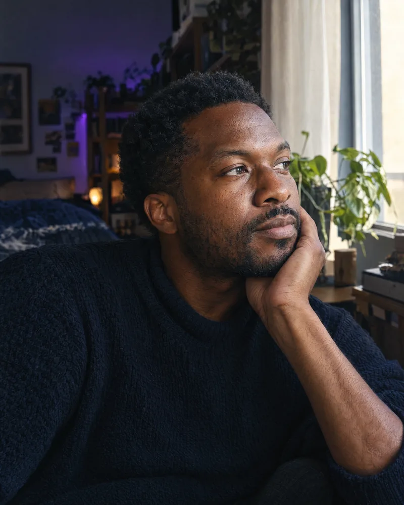 A man rests a hand on his jaw, looking toward the window.